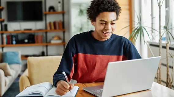 Schüler mit Schreibheft und Laptop beim Verfassen eines Berichts